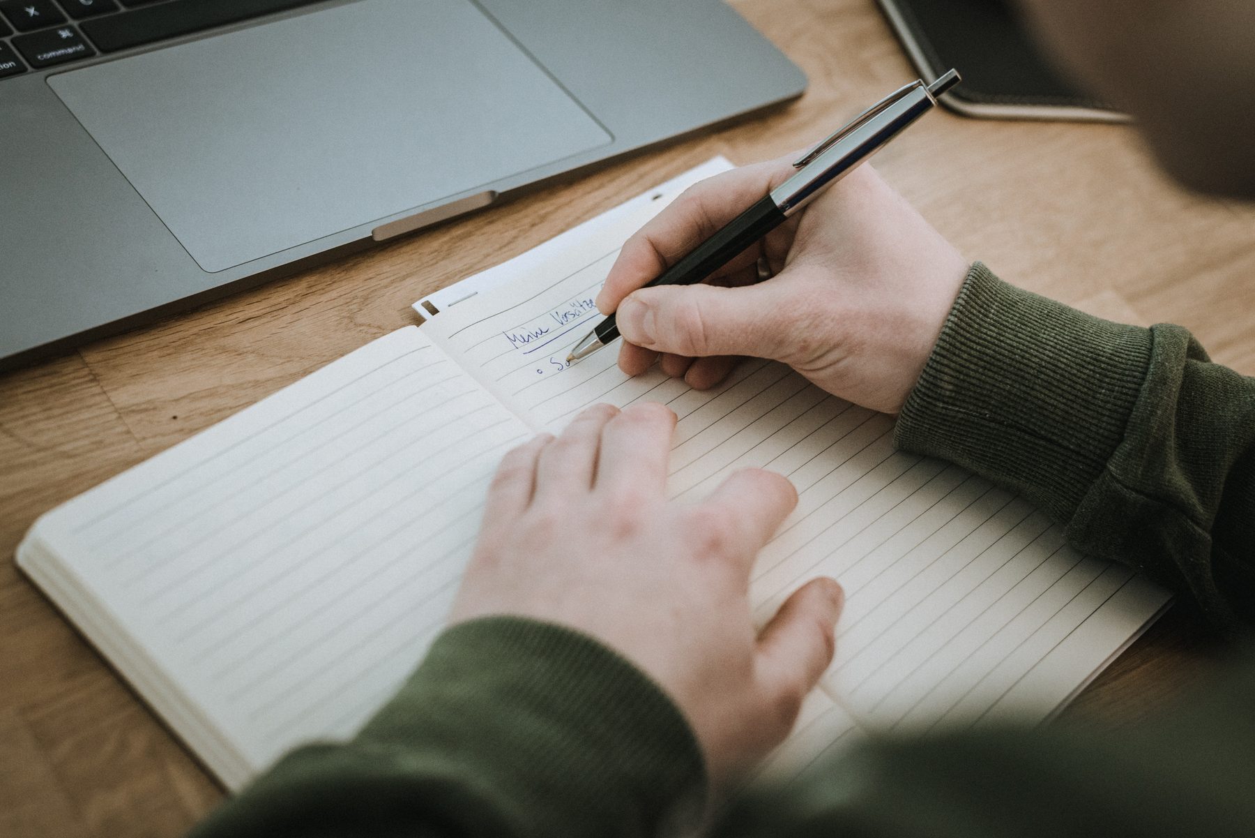 Hände halten einen Stift und notieren Gedanken in ein aufgeschlagenes Notizbuch neben einem Laptop